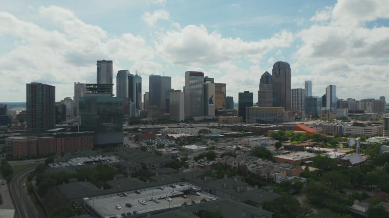 Panoramic aerial view of downtown skyscrapers behind rush highway. Forwards fly above low buildings. View against bright clouds in sky. Dallas, Texas, US