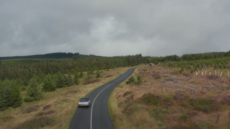 Following silver vintage sports car driving on curvy road in wooded landscape. Overcast sky on rainy day. Ireland