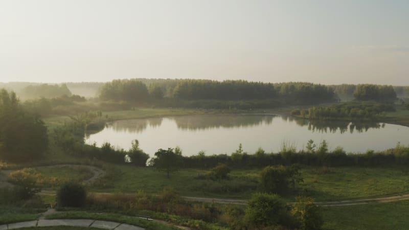 Aerial shot of a still lake with reflections of the landscape and animals grazing under soft sunrise golden light.