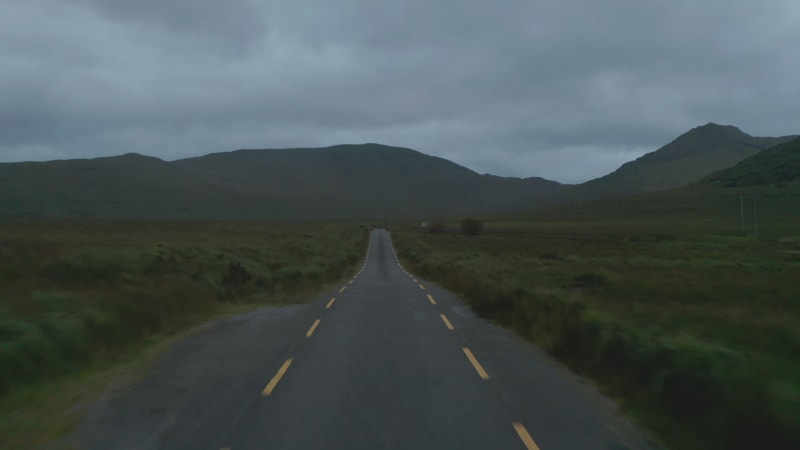 Forwards fly above narrow country road leading on old stone bridge over stream. Mountains against cloudy sky in background. Ireland