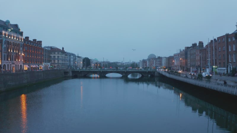 Low flight above calm Liffey river at dusk. Pedestrians walking on bridge. Reflections on water surface. Dublin, Ireland