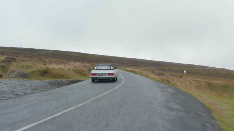 Forwards flyover historic sports car driving on curvy road in autumn countryside. Grasslands under overcast sky. Ireland