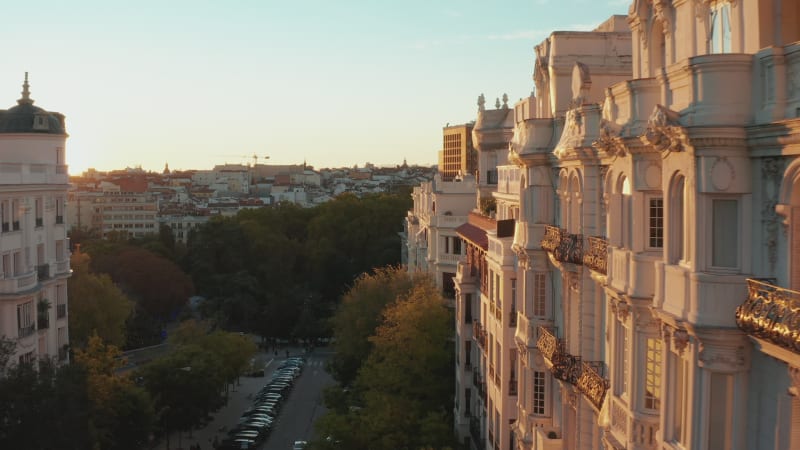 Forwards fly above wide street and along beautiful decorative facade of historic multistorey palace lit by bright sunset light.