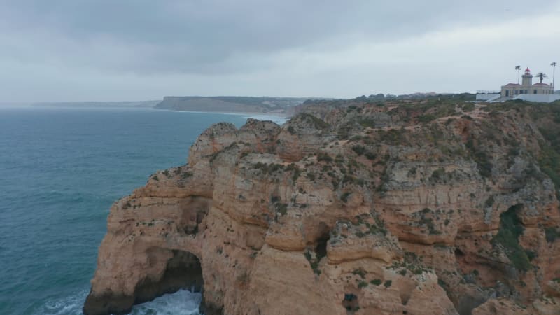 Aerial drone flying forward near rocky coloured cliffs in Lagos, Portugal, along coast with lighthouse on background, cloudy day