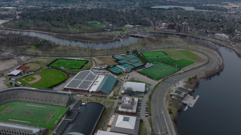 Aerial panoramic view of multilane road winding on Charles river waterfront around sports area. Tilt up reveal of residential suburbs. Boston, USA