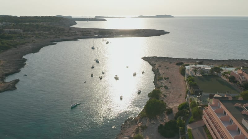 Aerial view of boats parked in a silent and calm sea with empty resort at shore with a football ground and greenery in Ibiza in Spain