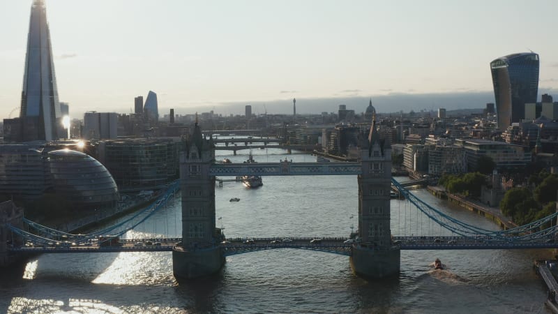 Aerial view of heavy traffic on Tower Bridge across River Thames. Backwards reveal of water surface reflecting sunshine. London, UK