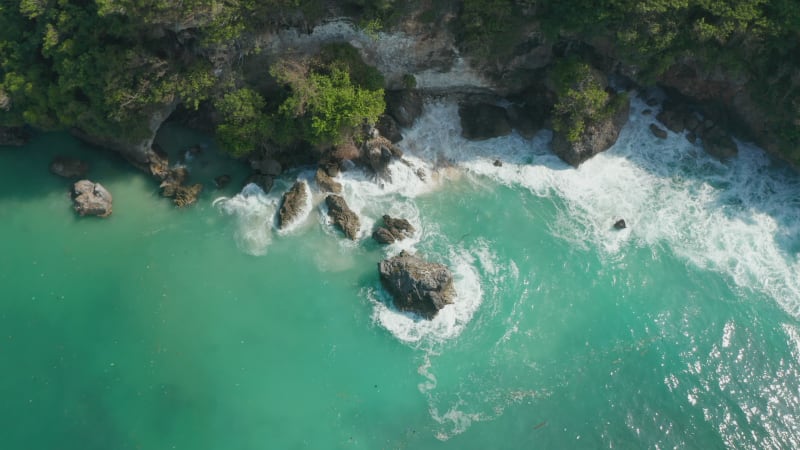 Blue waves crashing on the tropical coastline. Aerial tilting view of turquoise blue water foaming around the rocky cliffs