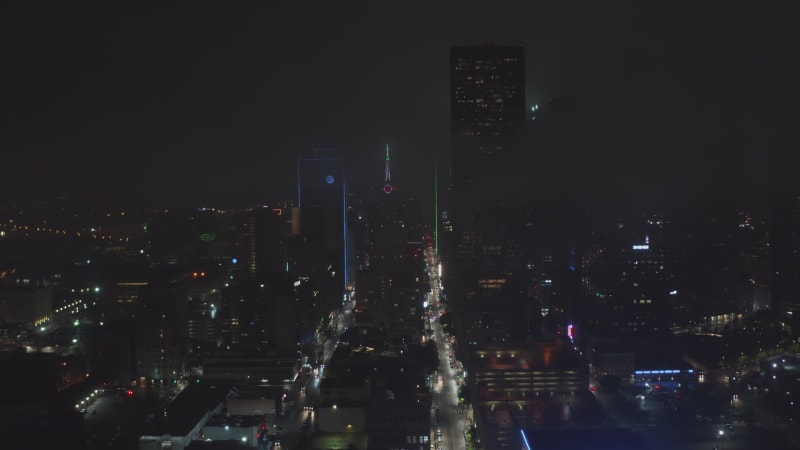 Drone flying over illuminated straight street between tall buildings around Mercantile National Bank Building with tower and big neon clock. Night aerial view of downtown. Dallas, Texas, US