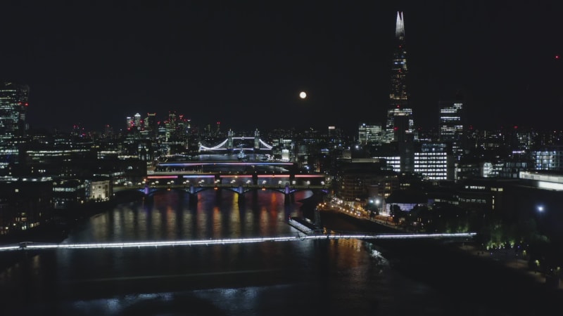 Fly above River Thames at Millennium Footbridge. Aerial view of illuminated buildings and landmark at night. London, UK