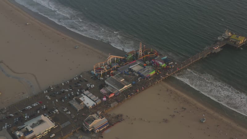 Flying away from Santa Monica Pier, Los Angeles at beautiful Sunset with Tourists, Pedestrians walking having fun at theme park rollercoaster with ocean view waves crashing