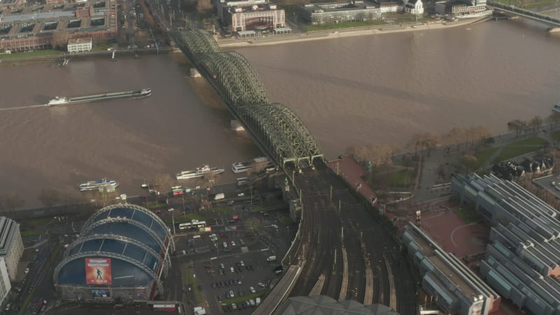 Aerial footage of cargo boat passing on wide Rhine river near railway bridge. Heavy traffic on waterfront road. Cologne, Germany