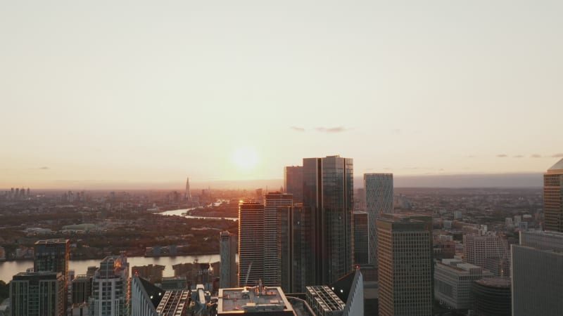 Fly over top of futuristic tall building. Elevated view of group of skyscrapers in Canary Wharf borough. Cityscape against sunset sky. London, UK