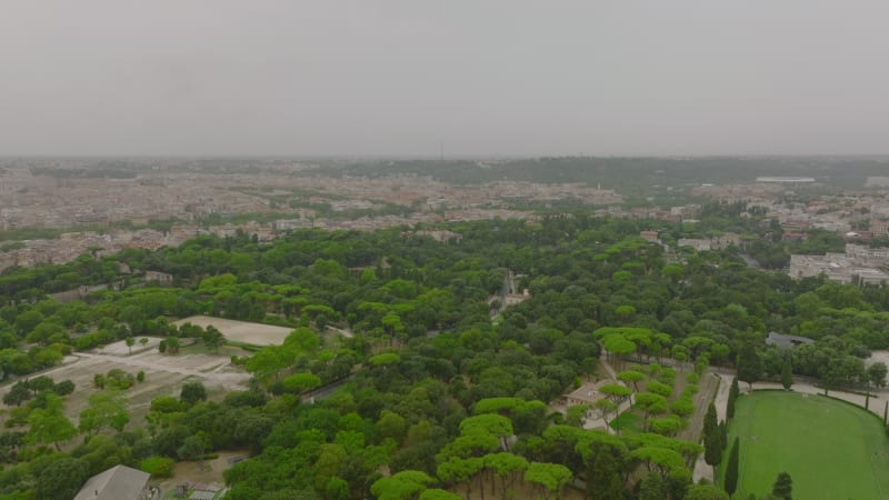 Aerial panoramic view of large city on cloudy day. Forwards fly above park with green trees and equestrian complex. Rome, Italy