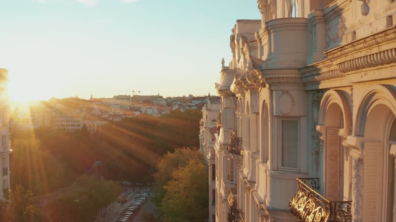 Forwards fly along historic palace with decorated facade. Elevated view of buildings in city. Illuminated by bright setting sun.