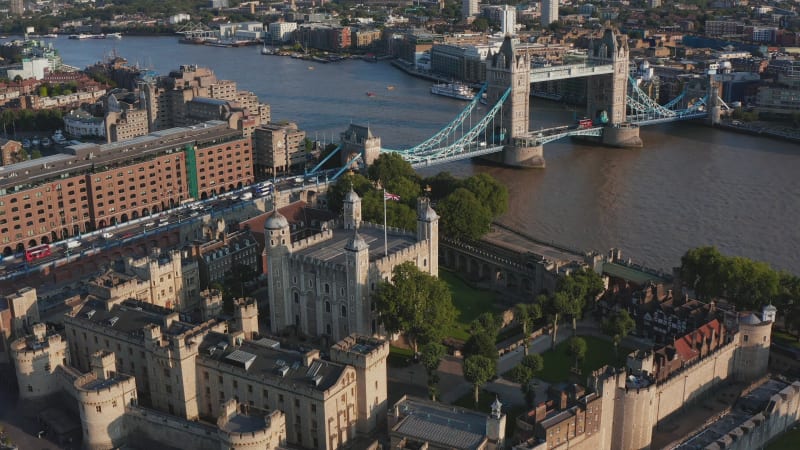 Aerial view of medieval royal castle Tower of London and Tower Bridge spanning River Thames. Tilt up reveal of buildings on other bank. London, UK