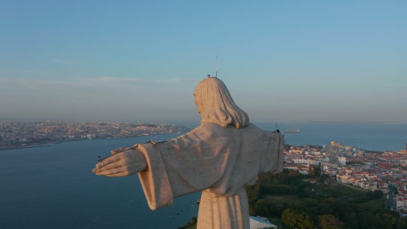 Big stone statue of Jesus illuminated by last sun rays of day. Drone camera flying behind Christ the King Sanctuary in Almada.  Lisbon, capital of Portugal.