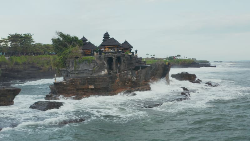 Closing in on empty Tanah Lot temple on dark dangerous cliffs in Bali, Indonesia. Famous Hindu temple on rocky cliff off the shore being pounded by strong ocean waves in bad weather