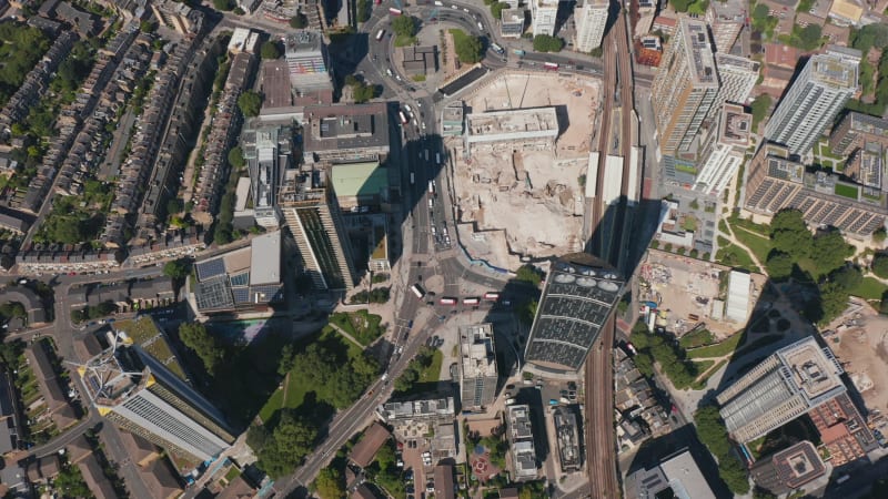 Tilt down aerial footage of traffic on road between tall modern buildings at Elephant and Castle. Construction site in city on brownfield between multilane road and railway track. London, UK