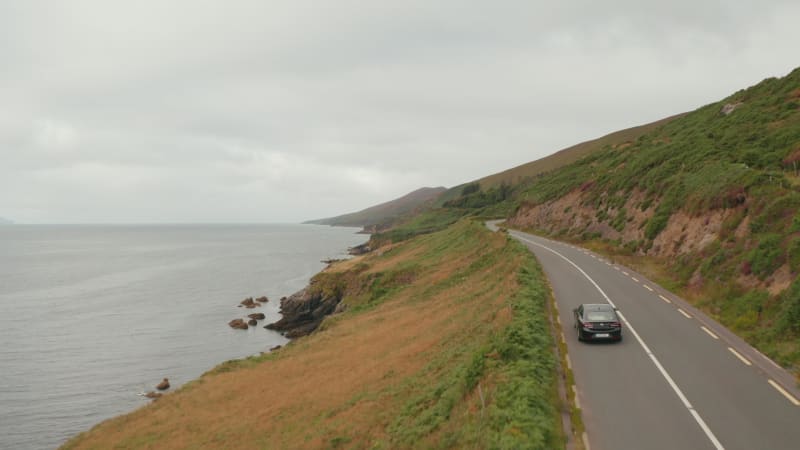 Forwards tracking of car driving on road winding above sea coast. Meadows and pastures in slope above rocky cliffs. Ireland