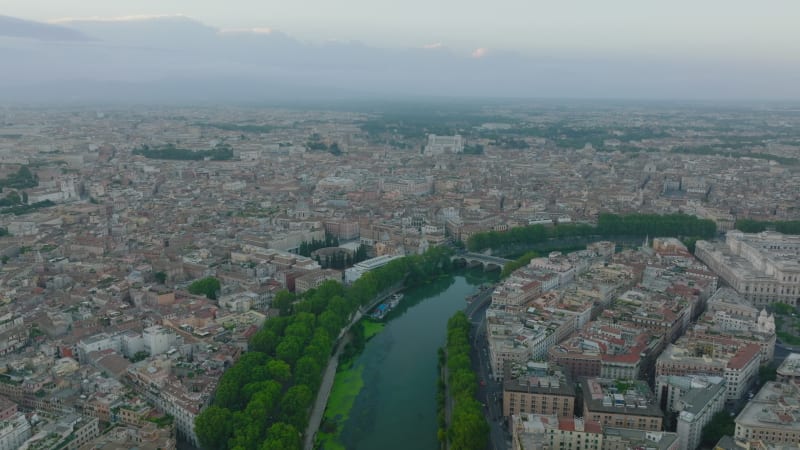 Aerial footage of Tiber river bending through historic city centre. Rows of lush green trees on riverbanks. Rome, Italy