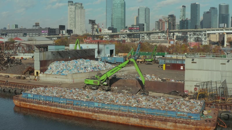 Aerial ascending footage of green loader standing on waterfront near barge loaded with plastic waste. Recycling facility. New York City, USA