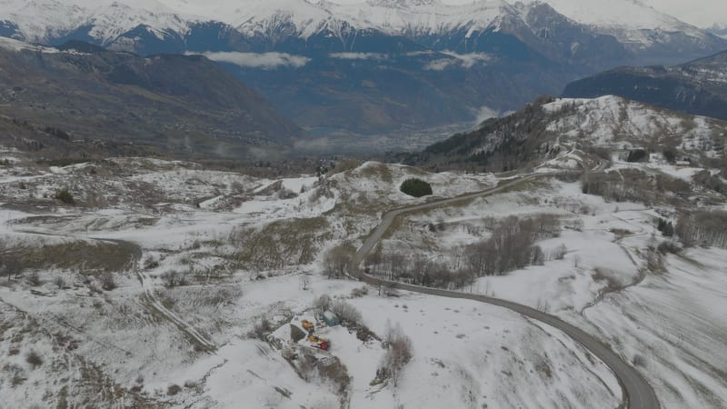 Forwards tilt down shot over an empty curved mountain road in snow covered landscape in Les Sybelles, France in cloudy weather