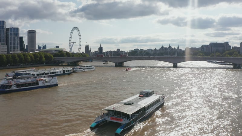 Forwards tracking of ship floating on glittering water surface of River Thames. View against sunshine. Silhouette of London Eye and Houses of Parliament over bright clouds in sky. London, UK