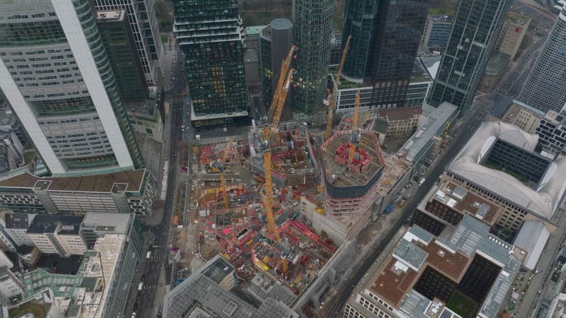 High angle shot of construction site of new building. Orange tower cranes and machinery. Tilt up reveal of downtown skyscrapers. Frankfurt am Main, Germany