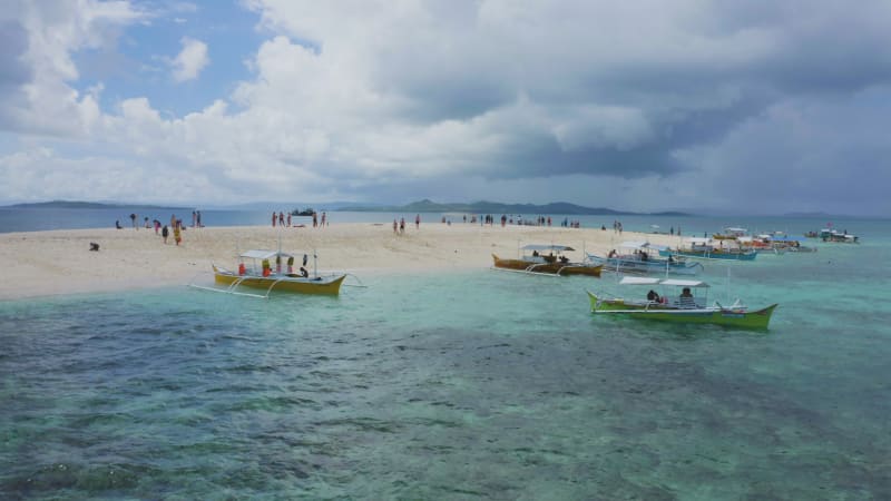 Low approach over crystal blue water to a sandbar with beachgoers and boats