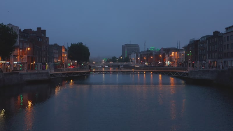 Pedestrians walking on footbridge across river in evening city. Ascending footage of town after sunset. Dublin, Ireland