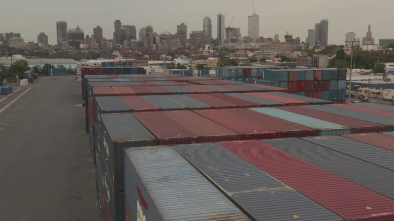 Flying over Red and Blue industrial cargo containers in docks with New York City with skyline in background on cloudy day