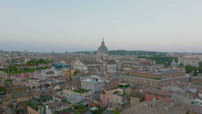 Forwards fly above old apartment houses with green plants on rooftop terraces. Heading towards dome at San Carlo al Corso basilica. Rome, Italy