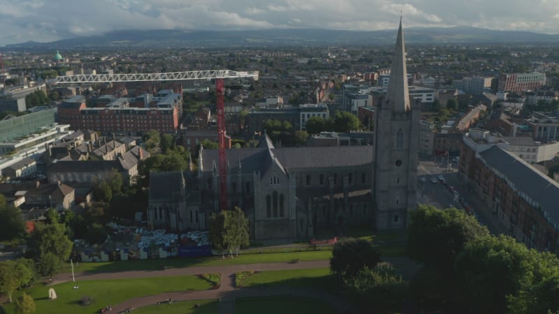 Slide and pan footage of old stone church surrounding park and town in background. Red and white tower crane. Dublin, Ireland