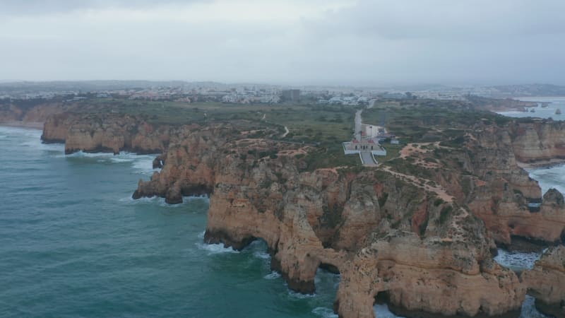 Amazing aerial drone view of Lagos, Portugal rocky coastline with lighthouse, cloudy grey day, circle pan