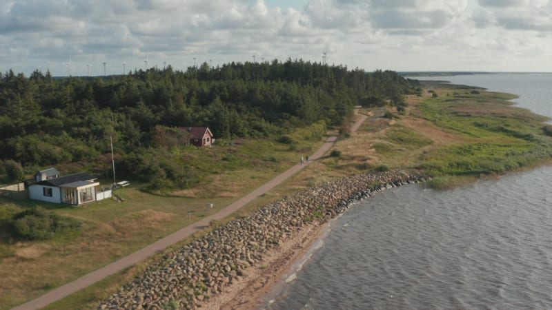 Family walking on pathway along sea coast. Aerial footage of coastal landscape with trees and wind turbines in background. Denmark