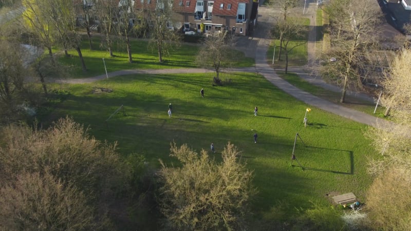Birdseye downwards tilt drone shot to reveal people playing football at a shaded park on a sunny day in Houten, Netherlands.