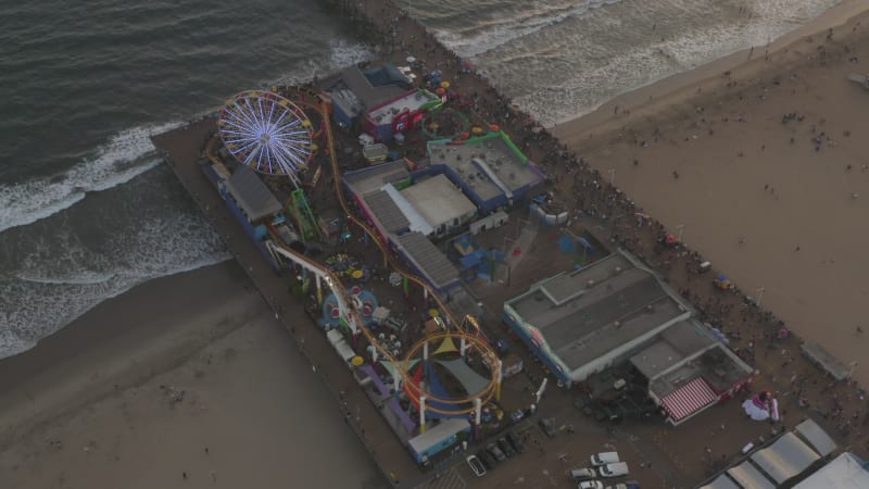 Circling Santa Monica Pier, Los Angeles from above at beautiful Sunset with Tourists, Pedestrians having fun at theme park Ferris Wheel with ocean view waves crashing