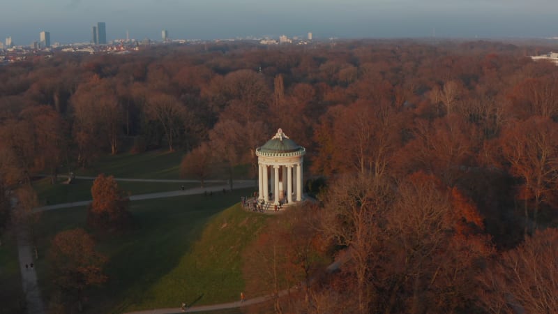 Epic Scenic Aerial Slide circle around The Monopteros in English Garden in Munich, Germany surrounded by beautiful Orange Trees in Public Park with Green Grass, Aerial Dolly slide right