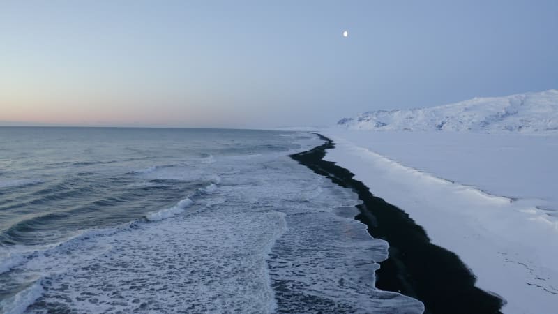 Flying over Black Beach with white arctic snow mountains in background in Iceland in Winter Snow, Ice, Waves, Water