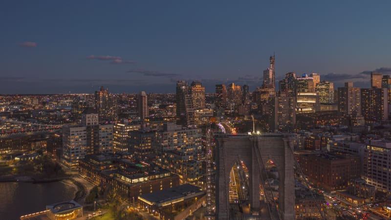 Forwards fly above Brooklyn bridge at twilight. Dusk hyperlapse of heavy traffic on roads and lighted windows on buildings. Brooklyn, New York City, USA