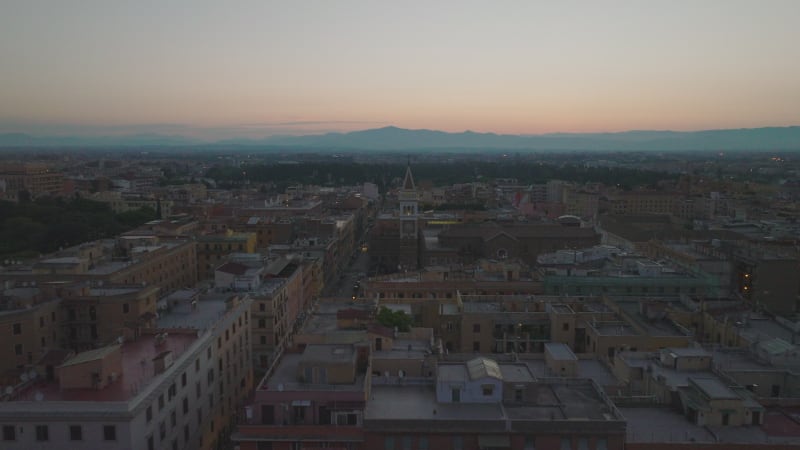 Backwards fly above buildings in urban borough at dawn. Silhouette of mountain ridge against colour sky. Rome, Italy