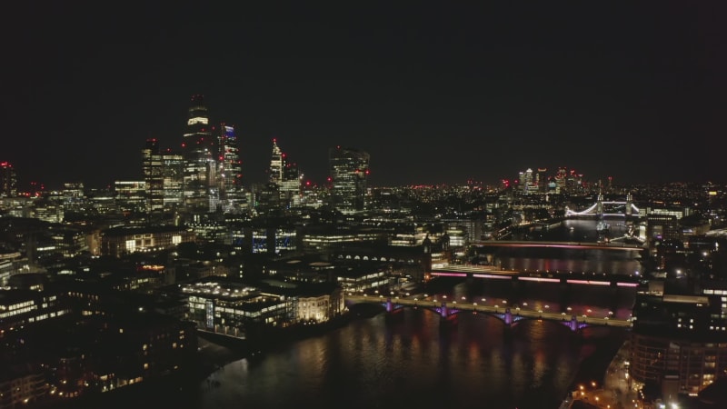 Night scene of large town. Modern tall buildings on City financial and economical centre. Backwards reveal of Thames river and its bridges. London, UK