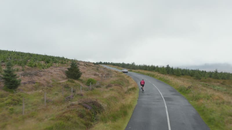 Cyclists and cars on mountain road leading up hill. Forest with young trees along road. Overcast sky. Ireland