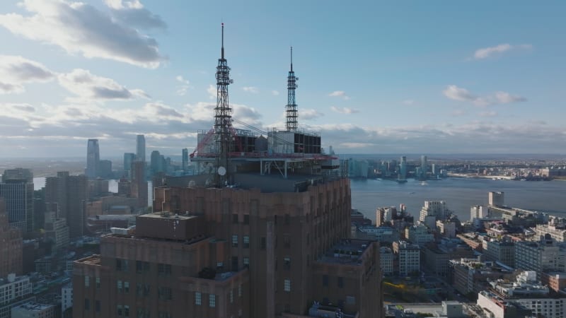 Orbit shot around top of tall building with steel construction and spires with antennas. Downtown skyline in background. Manhattan, New York City, USA