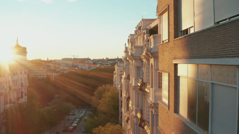 Elevated forwards fly along row of buildings in city centre.  Old palace with decorated facade illuminated by bright setting sun.