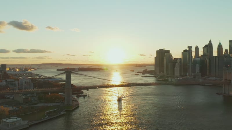 Aerial panoramic view of Brooklyn Bridge against sunset. Cruise ship floating of glowing water surface. Skyscrapers on waterfront. Manhattan, New York City, USA