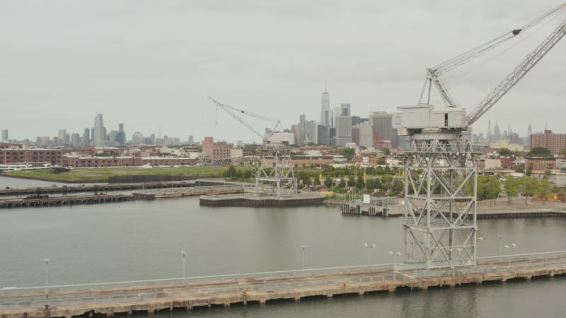 Flying in between industrial cranes in docks with New York City skyline in background and river