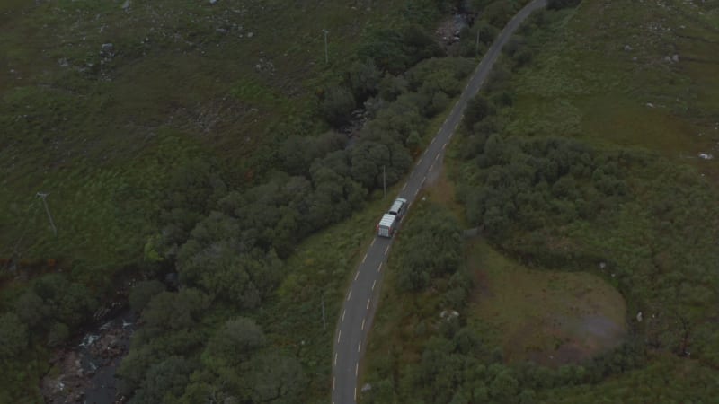 Forwards tracking of car with horse trailer driving on country road along stream. High angle view of valley with trees and shrubs. Ireland