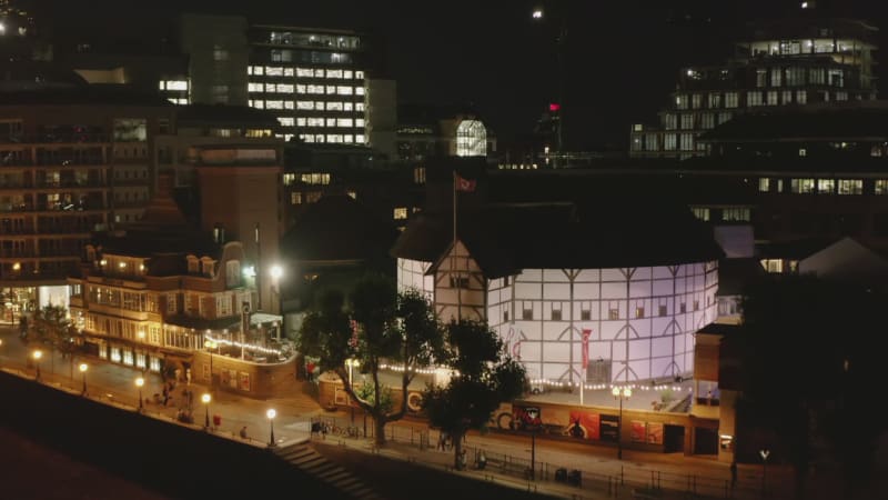 Forwards fly to illuminated waterfront. Aerial night view of buildings in city. People walking on street. London, UK